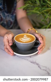 Green Leafy Background With A Close Up Photo Of Woman's Hands Holding A Light Blue Cup Of Coffee With Latte Art