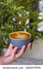 Green Leafy Background With A Close Up Photo Of Woman's Hands Holding A Light Blue Cup Of Coffee With Latte Art