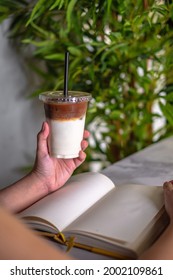 Green Leafy Background With A Close Up Photo Of Woman's Hands Holding A Cup Of Iced Spanish Latte While Reading A Book