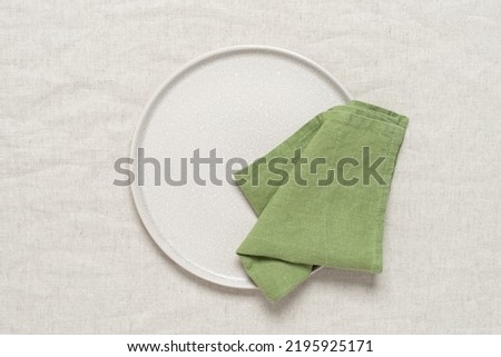 Green linen napkin on a clean beige plate, beige textile background. Top view, flat lay.
