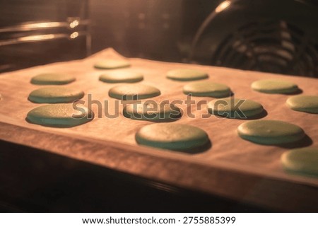 Green macaron shells baking on parchment paper inside oven, warm light and shallow depth of field, pastry process and homemade dessert preparation.