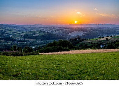 Green Meadow On The Hill At Sunset, Mountains And Forest In The Background. No People, Clear Sky, Few Buildings In The Background