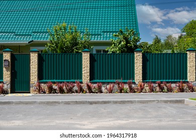 Green metal corrugated fence with brick pillars on the background of a beautiful house.