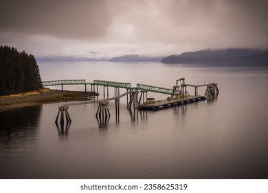  A green metal pier that extends into the calm water of Icy Strait Point, Alaska. The pier has a wooden deck and a small crane on it. - Powered by Shutterstock