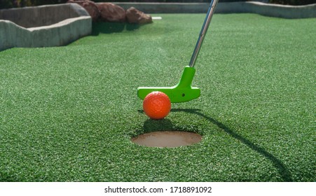 Green mini golf putter ready to tap an orange ball into the hole on green turf