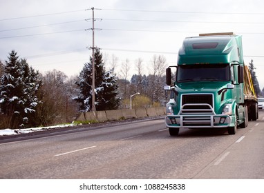 Green Modern Big Rig Semi Truck With Grille Protection Bumper Transporting Commercial Cargo Tightened With Slings On Flat Bed Semi Trailer On The Winter Snowy Road With Trees On Background