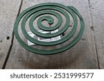 A green mosquito coil is placed on a wooden surface. The coil is still intact, and a metal holder is placed beside it.