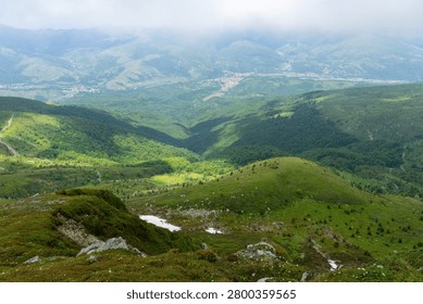 Green mountain landscape with sparse trees and distant peaks under a vibrant blue sky