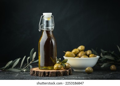 Aceitunas verdes en un bol blanco al lado de una botella con aceite de oliva y hojas en un fondo negro. Botella de aceite prensado frío. Cocina tradicional griega e italiana.