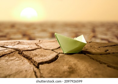 Green Paper Boat On Dry Cracked Earth With Summer Sunset Metaphor Climate Change, Global Warming And Drought.