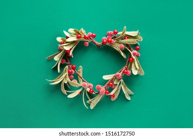 Green paper flat lay with decorative mistletoe with frosted leaves and red berries shaped as a heart