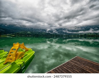 A green pedal boat with yellow slide is docked on Lac d'Aiguebelette in France, with misty mountains in the backdrop - Powered by Shutterstock