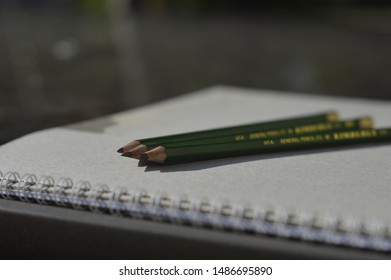 Green Pencils On Top Of A Sketchbook, Outside, On A Glass Table