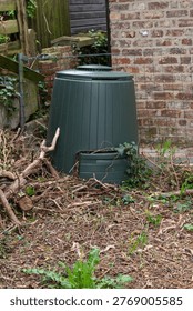 Green plastic compost bin in a backyard garden for organic waste recycling.Garden composter placed near a brick wall for making natural fertilizer.Sustainable gardening: composting organic kitchen and garden waste in a plastic container.Eco-friendly waste management: dark green compost tumbler in a home garden.Compost heap with a modern plastic bin for soil enrichment and ecological living