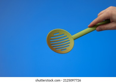 Green Plastic Heat-resistant Perforated Strainer Ladle Held By Caucasian Male Hand. Close Up Studio Shot, Isolated On Blue Background.