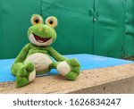 Green plush toy frog with big yellow eyes, forgotten by children on a wooden board, garage gates in the background, selective focus