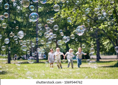 Green public park illuminated with sunbeams, colorful soap bubbles flying in air, group of little children playing active games