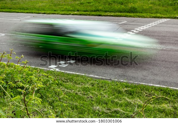 A green race\
car at speed crosses the finish lane of an asphalt track against a\
background of green grass and bushes. The car is blurred.\
Horizontal orientation. High quality\
photo