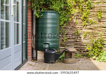A green rain barrel to collect rainwater and reusing it to water the plants and flowers in a backyard with a wattle fence made of willow branches on a sunny day