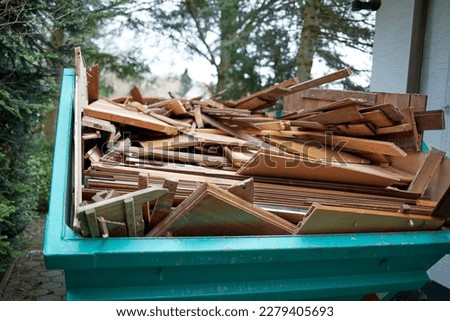 green recycling container filled with wood, next to a house