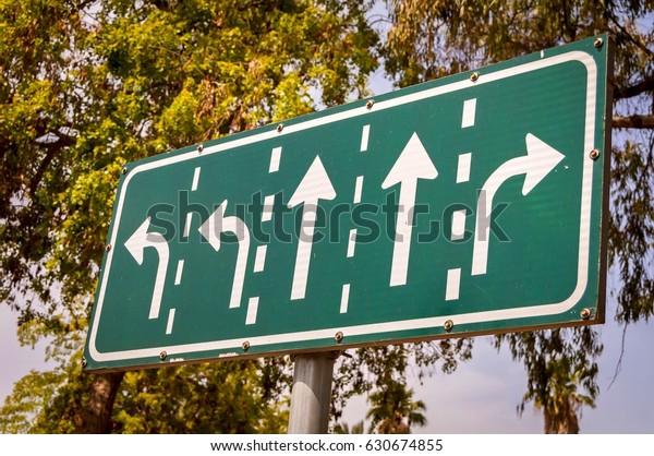 Green road\
intersection arrow sign with directions for five lanes on a sky and\
trees background stock\
image.