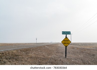 A Green Road Sign With The Name Of The Town Of Dagmar, Montana, Is Posted Over A Yellow Road Sign That Says, Rough Break, Posted Next To A Rural Country Road As Snow Blows Sideways In The Wind.