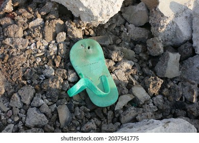 A Green Sandals Or Flip Flop Waste On A Beach Or Coast, Ruins Where There Are Some Rocks Around It. One Side Abandoned Broken Kid (small Size) Worn Out Slipper On Ground. As Missing Child Evidence.