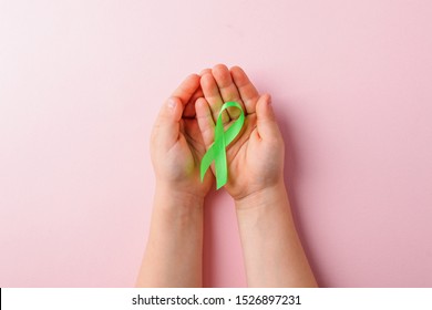 Green satin ribbon holding in kids hands on pink background. Liver, kidney cancer awareness, Glaucoma Awareness