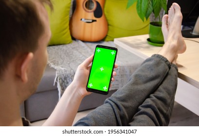 Green Screen And Chroma Key. Caucasian Man Holding Mobile Phone With Greenscreen Of Chromakey Mockup With Tracking Markers. Man Is Resting At Home And Watching Phone.