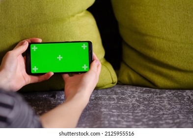 Green Screen And Chroma Key. Caucasian Woman Holding Mobile Phone With Greenscreen Of Chromakey Mockup With Tracking Markers. Woman Is Resting At Home And Watching Phone.