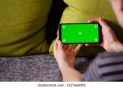 Green Screen And Chroma Key. Caucasian Woman Holding Mobile Phone With Greenscreen Of Chromakey Mockup With Tracking Markers. Woman Is Resting At Home And Watching Phone.