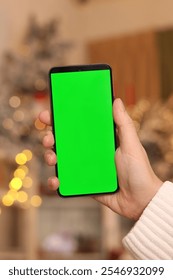 Green screen phone in a female hand. Gadget on the background of Christmas lights. Close-up of a female hand and a smartphone with a green screen, New Year. Modern mobile phone. Mockup. Technology