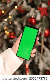 Green screen phone in a female hand. Gadget on the background of Christmas lights. Close-up of a female hand and a smartphone with a green screen, New Year. Modern mobile phone. Mockup. Technology