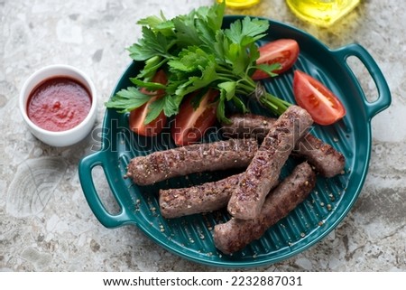 Green serving tray with grilled serbian cevapi or cevapcici sausages with fresh parsley, tomatoes and dip, elevated view on a brown granite background