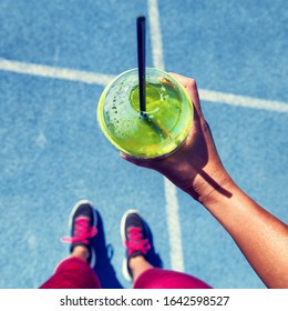 Green Smoothie Woman Drinking Plastic Cup Breakfast Meal Takeaway To Go After Morning Run On Blue Tracks. Healthy Lifestyle Sporty Person Pov Of Hand Holding Glass With Running Shoes Feet Selfie.