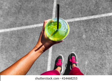 Green Smoothie Woman Drinking Plastic Cup Breakfast Meal Takeaway To Go After Morning Run On City Streets. Healthy Lifestyle Sporty Person Pov Of Hand Holding Glass With Running Shoes Feet Selfie.