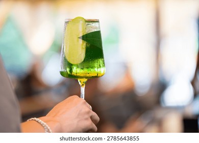 Green spritz cocktail with a slice of green apple and leaf in a wine glass held by a hand.