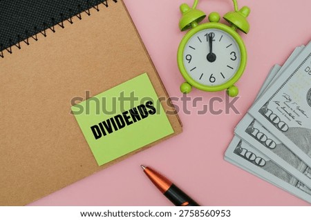 A green sticky note with text DIVIDENDS on a notebook, featuring a stack of US dollars, a pen, and an alarm against coloured background. Concept of passive income and time value of money