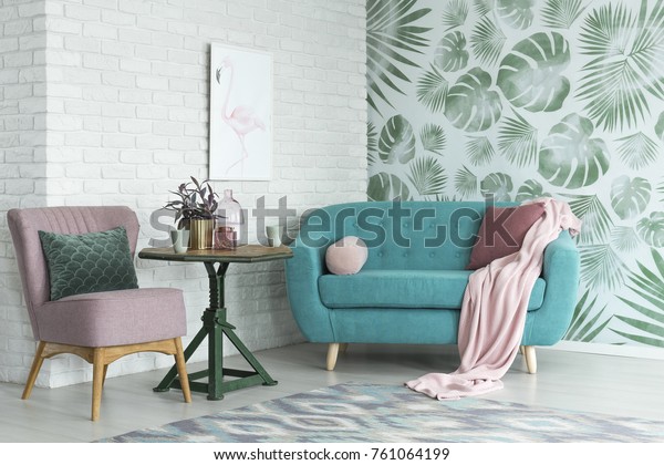 Green table with a plant between pink chair and blue sofa in floral living room with wallpaper and poster.