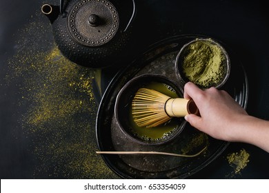 Green tea matcha powder and hot drink in black bowls standing with iron teapot, bamboo traditional tools spoon and whisk in hand in vintage tray over dark metal background. Top view with space