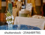 A green textured orthodox baptismal candle adorned with white flowers and greenery sits on an ornate blue table in a softly focused, elegant setting with muted background details.