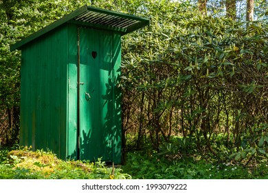 Green timber frame outhouse with heart shape hole in door. Rustic wooden latrine among green vegetation. Dry outside toilet close to non blooming rhododendron bush. Green wood jakes hided among bushes