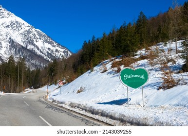Green Traffic Road Sign In Heart Shape.
Welcome In Styria. Beautiful Winter Landscape With Empty Country Road, Snow Capped Mountain And Fir Trees.
