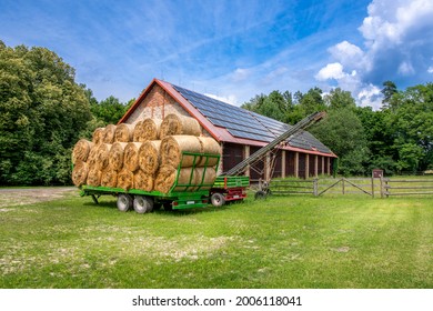 Green trailer filled with hay bales parked in the front of brick barn on a farm. Solar panels installed on the roof of the barn. 