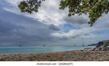 Green tree branches hang over the beach. Two chaise lounges stand on the sand. Yachts are visible on the calm turquoise ocean. Clouds in the blue sky. Seychelles. Mahe Island. Beau Vallon - Shutterstock ID 2202861875