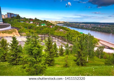 Green trees on the slope in front of the panorama of the city and the river flowing below on a bright sunny day. Multicolored private houses. Park of Salavat Yulayev, Ufa, Bashkortostan, Russia.