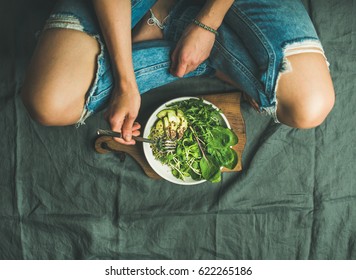 Green vegan breakfast meal in bowl with spinach, arugula, avocado, seeds and sprouts. Girl in jeans holding fork with knees and hands visible, top view, copy space. Clean eating, detox food concept - Shutterstock ID 622265186