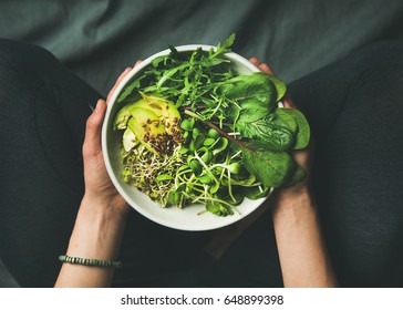 Зеленый веганский завтрак в миске со шпинатом, рукколой, авокадо, семенами и ростками. Девушка в леггинсах держит тарелку с видимыми руками, вид сверху. Чистое питание, диета, концепция веганского питания