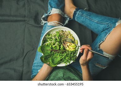 Green vegan breakfast meal in bowl with spinach, arugula, avocado, seeds and sprouts. Girl in jeans holding fork with knees and hands visible, top view, copy space. Clean eating, vegan food concept