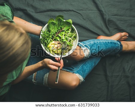 Green vegan breakfast meal in bowl with spinach, arugula, avocado, seeds and sprouts. Girl in jeans holding fork with knees and hands visible, top view. Clean eating, detox, vegetarian food concept